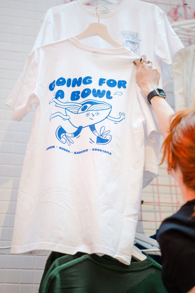 A white and blue House of Fu graphic tee from our merchandise collection. The graphic print reads 'Going for a Bowl' and 'Ramen, Gyoza, Karaoke, Cocktails' with a cartoon, walking Ramen bowl illustration.